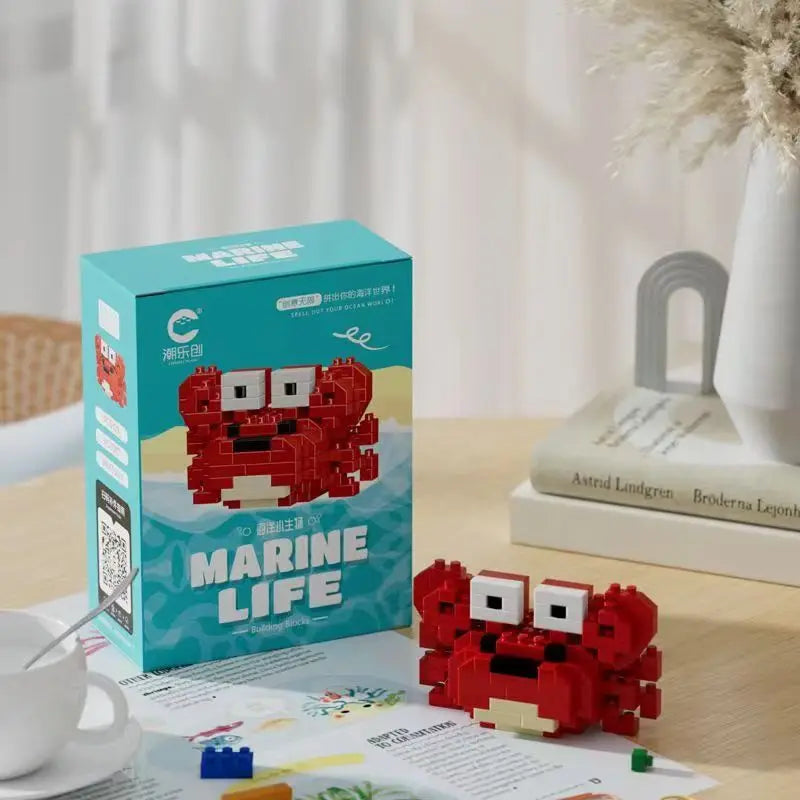 Bloco de montar em formato de caranguejo marinho, feito com blocos de brinquedo vermelho, branco e pretos, apresentado ao lado da caixa do produto "Marine Life" em uma mesa de madeira.