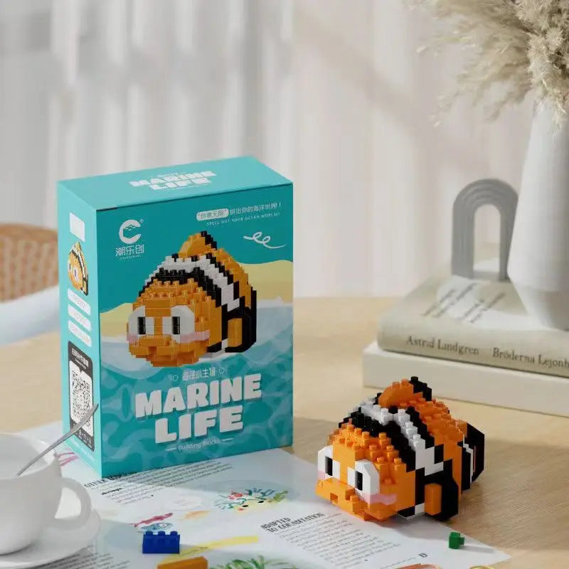 Imagem de um brinquedo de peixe-palhaço Nemo construído com blocos de montar, com cores laranja, branco e preto, sobre uma mesa de madeira ao lado de sua embalagem que tem a palavra 'Marine Life'.