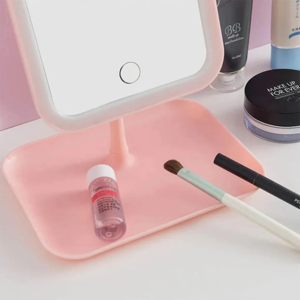 Espelho LED de maquiagem com base retangular, pincel, produtos de beleza e itens de cuidados faciais em fundo rosa, ideal para rotina de beleza e preparo de maquiagem.