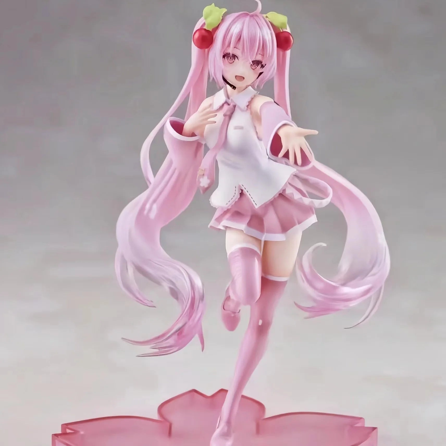 Figura de anime Hatsune Miku Sakura com cabelo rosa e saia rosa, vestida com roupa escolar, em pose dinâmica. Ela possui penteados com acessórios vermelhos e verdes, com longas tiras de cabelo e expressão alegre.