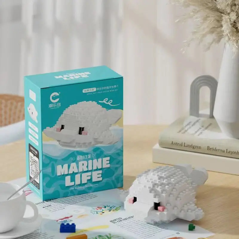 Boneco de construção em formato de baleia branca feito com blocos de montar, com tema marinho, em uma mesa com livros e uma xícara ao fundo.